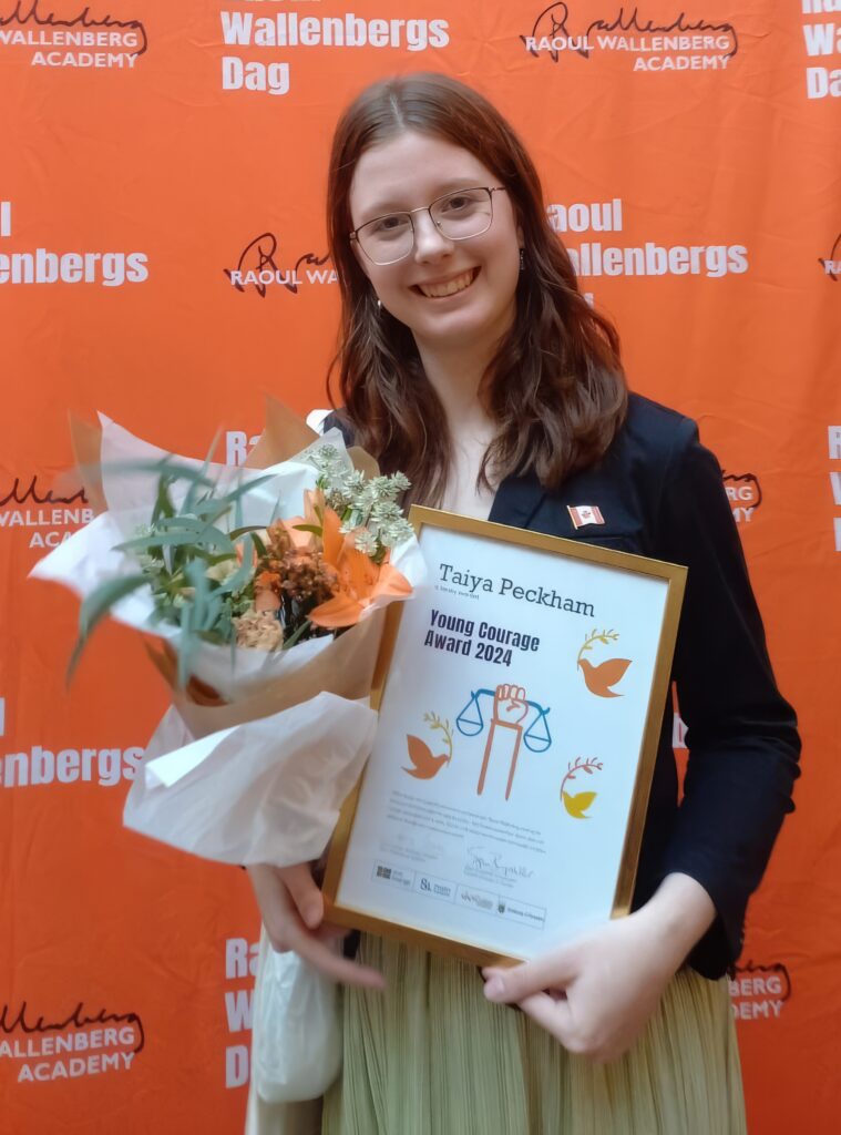 Taiya Peckham smiling towards the camera. She is holding her award and a bouquet of flowers, standing against an orange background.