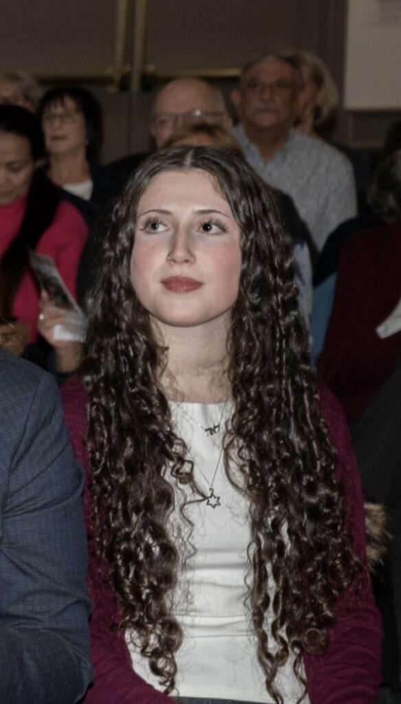 Leah is looking away from the camera. She is wearing a white shirt and a maroon sweater, and an audience is in the background.